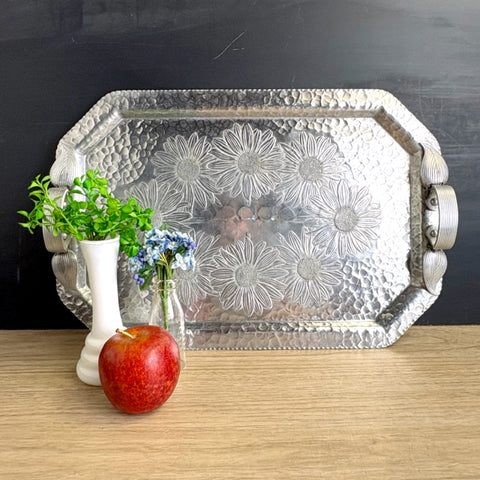 Decorative metal tray with floral patterns on a wooden surface, next to an apple and small vase with flowers.