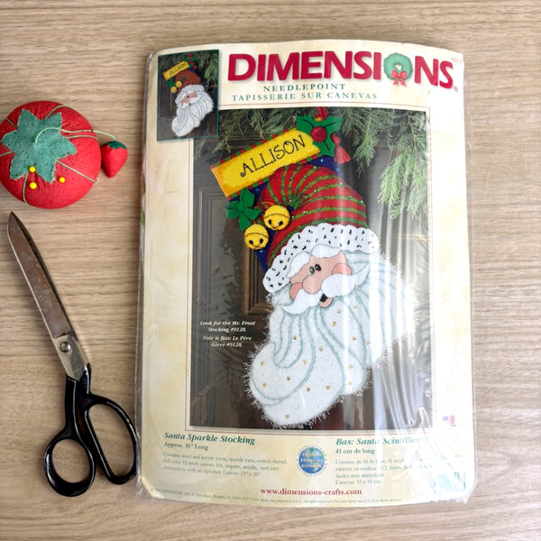 Dimensions needlepoint kit packaging with a Santa design, scissors, and a red and green fabric item on a wooden surface.