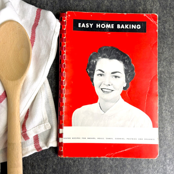 Vintage 'Easy Home Baking' cookbook with a woman's portrait on a red background, next to a wooden spoon and kitchen towel.