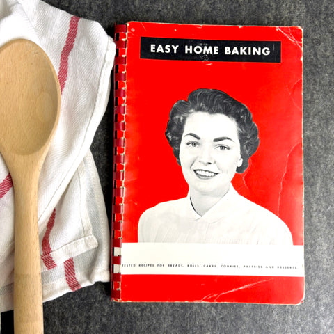 Vintage 'Easy Home Baking' cookbook with a woman's portrait on a red background, next to a wooden spoon and kitchen towel.