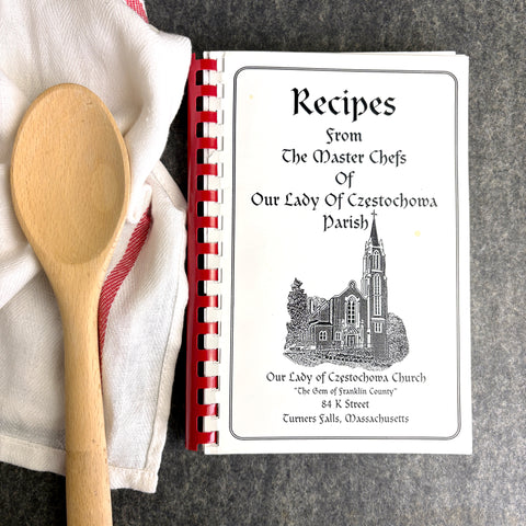 Cookbook titled 'Recipes from The Master Chefs Of Our Lady Of Czestochowa Parish' with a wooden spoon and towel on a gray surface.
