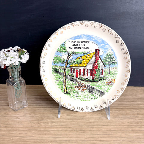 Decorative plate with a house illustration and text on a stand next to a vase with flowers against a dark background.